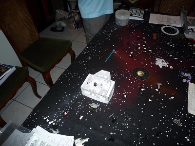 Overhead view of a 3D cardboard model of the Beowulf's bridge, next to a Beowulf starship miniature, pulled back far enought to a large part of the surrounding starfield battlemap